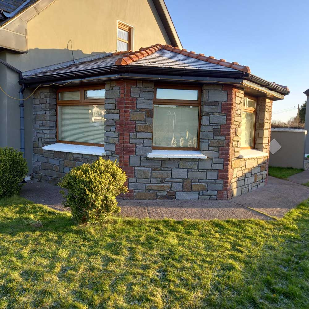 Stone facade with windows, brick accents, and a tiled roof, extending from a beige house, in a sunny yard.