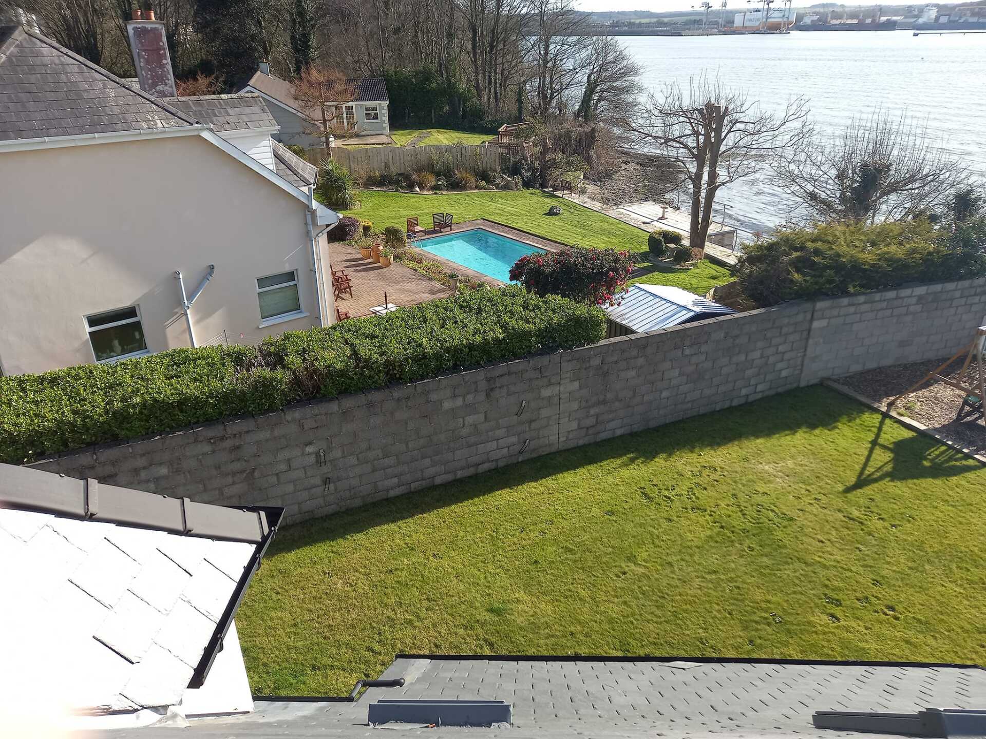Lush green lawn with a low wall bordering a yard with a pool, leading to a water view. Sunny day.