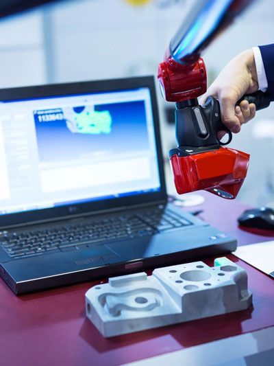 Handheld 3D scanner capturing a machined part with live mesh on laptop.