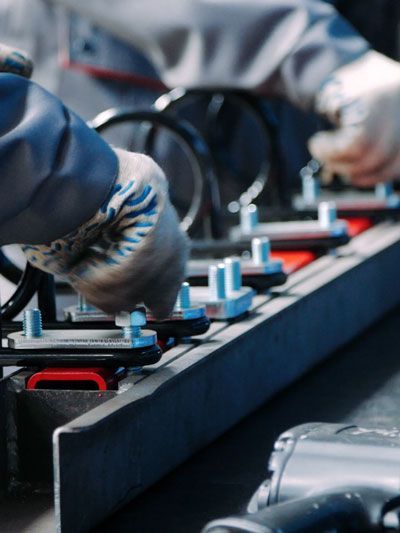 Workers assemble parts on an assembly line, wearing gloves and grey coveralls.