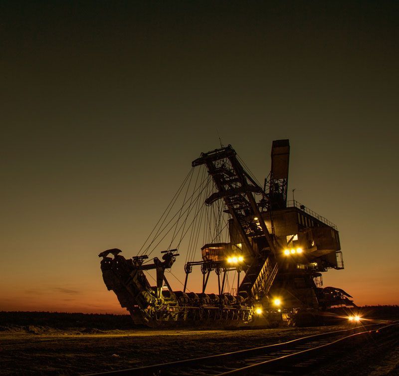 A giant excavator at dusk with lights illuminated, next to train tracks.