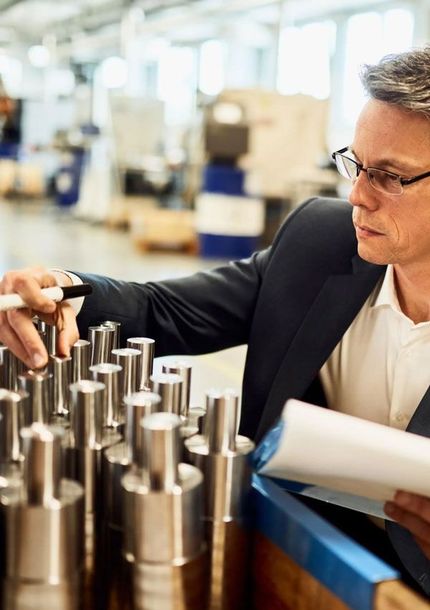 Man in glasses inspecting metal parts in a factory, holding a pen and a clipboard.