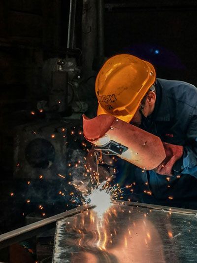 Welder wearing a yellow hard hat and welding a metal frame; sparks fly in a dimly lit workshop.