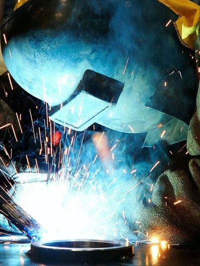 Welder in action, wearing a helmet, sparks flying, creating a bright blue arc on metal.