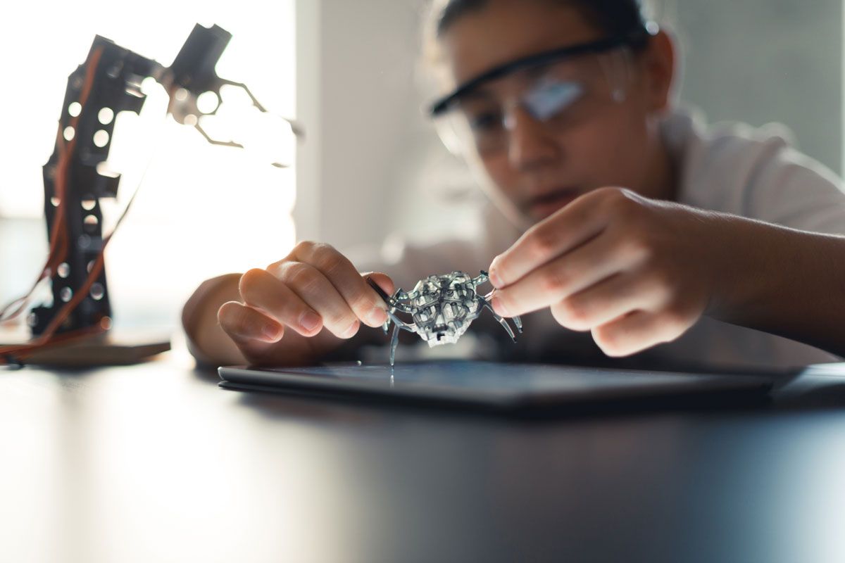 A person with safety glasses examines a circuit board while working on a robotic arm.