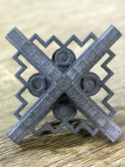 Gray, decorative, cross-shaped object with a zig-zag frame and stylized center on a wooden surface.