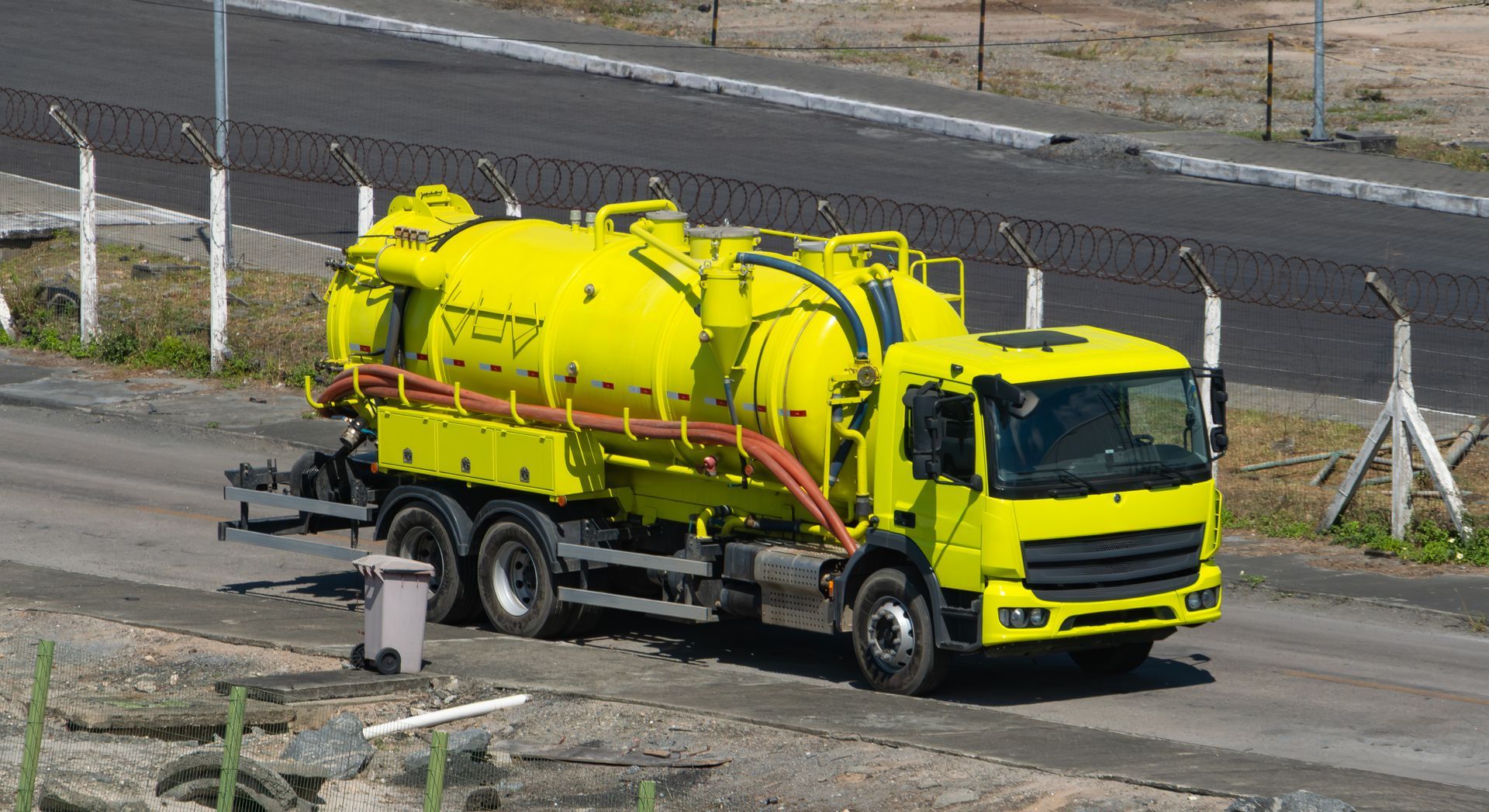 Yellow vacuum truck on road next to a fence.