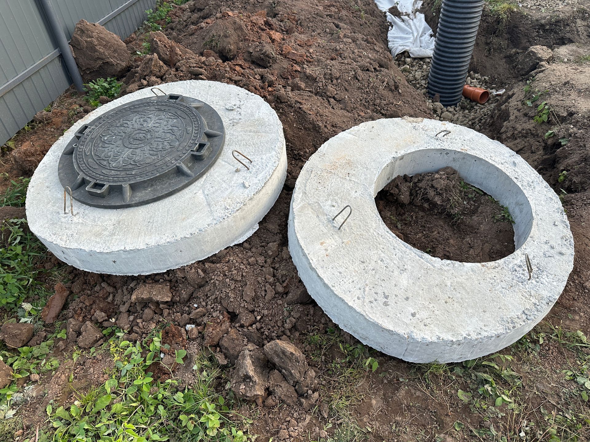 Two concrete rings with a manhole cover on top, lying on soil and grass.