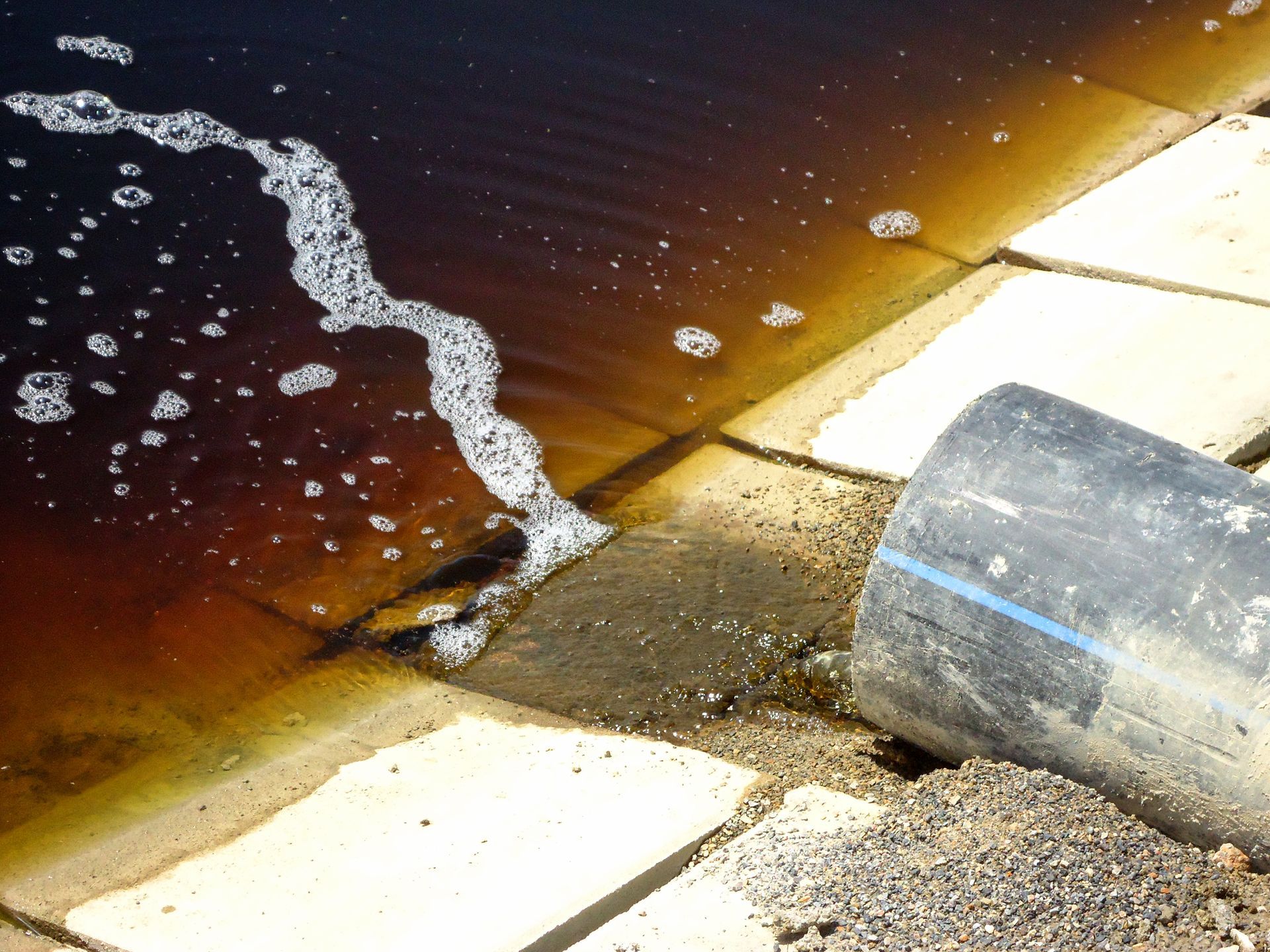 Dark, foamy water flows from a pipe onto tiled concrete. The water is brown and the tiles are light-colored.
