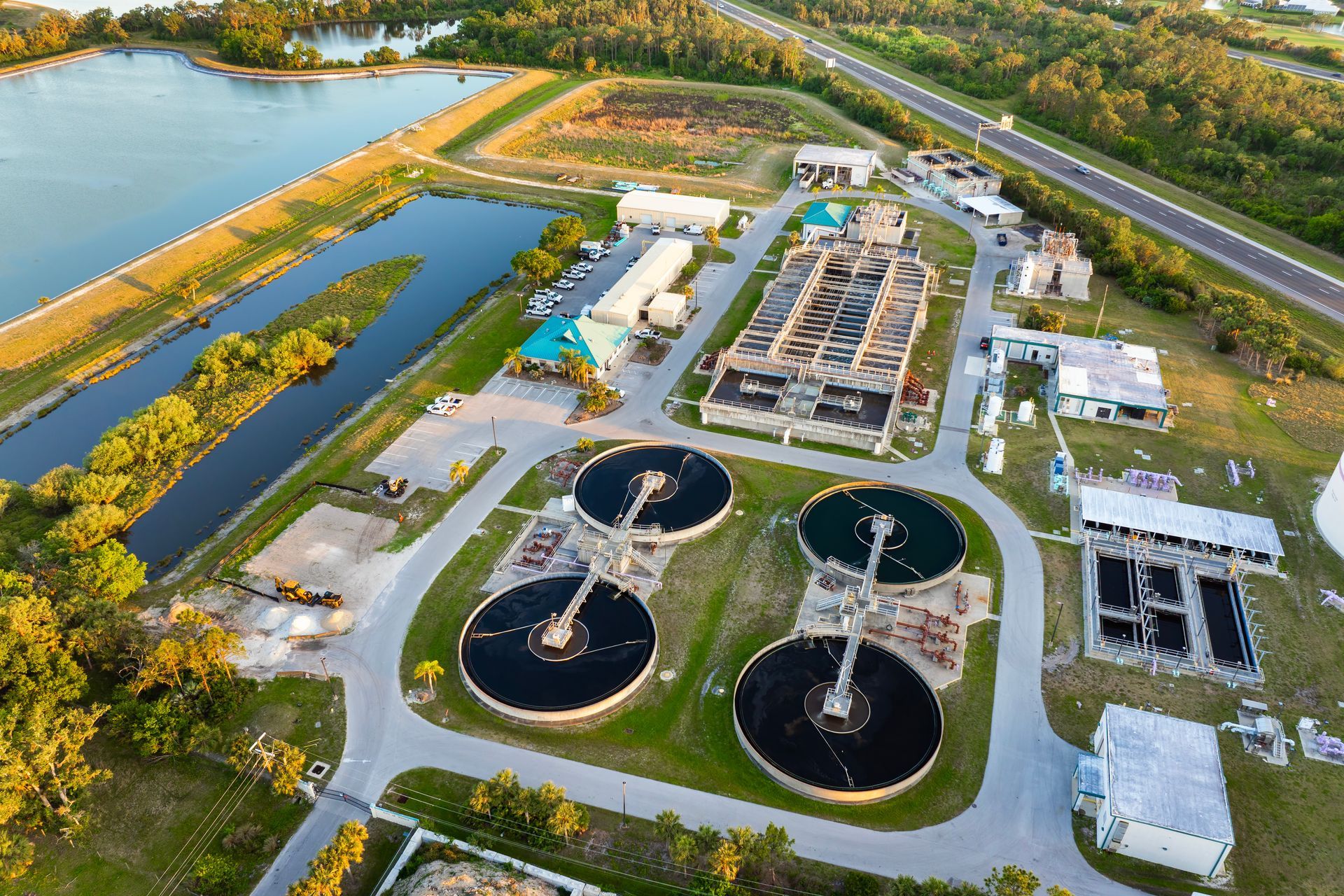 Aerial view of a wastewater treatment plant with several circular tanks and rectangular structures. Green and blue water features are nearby.