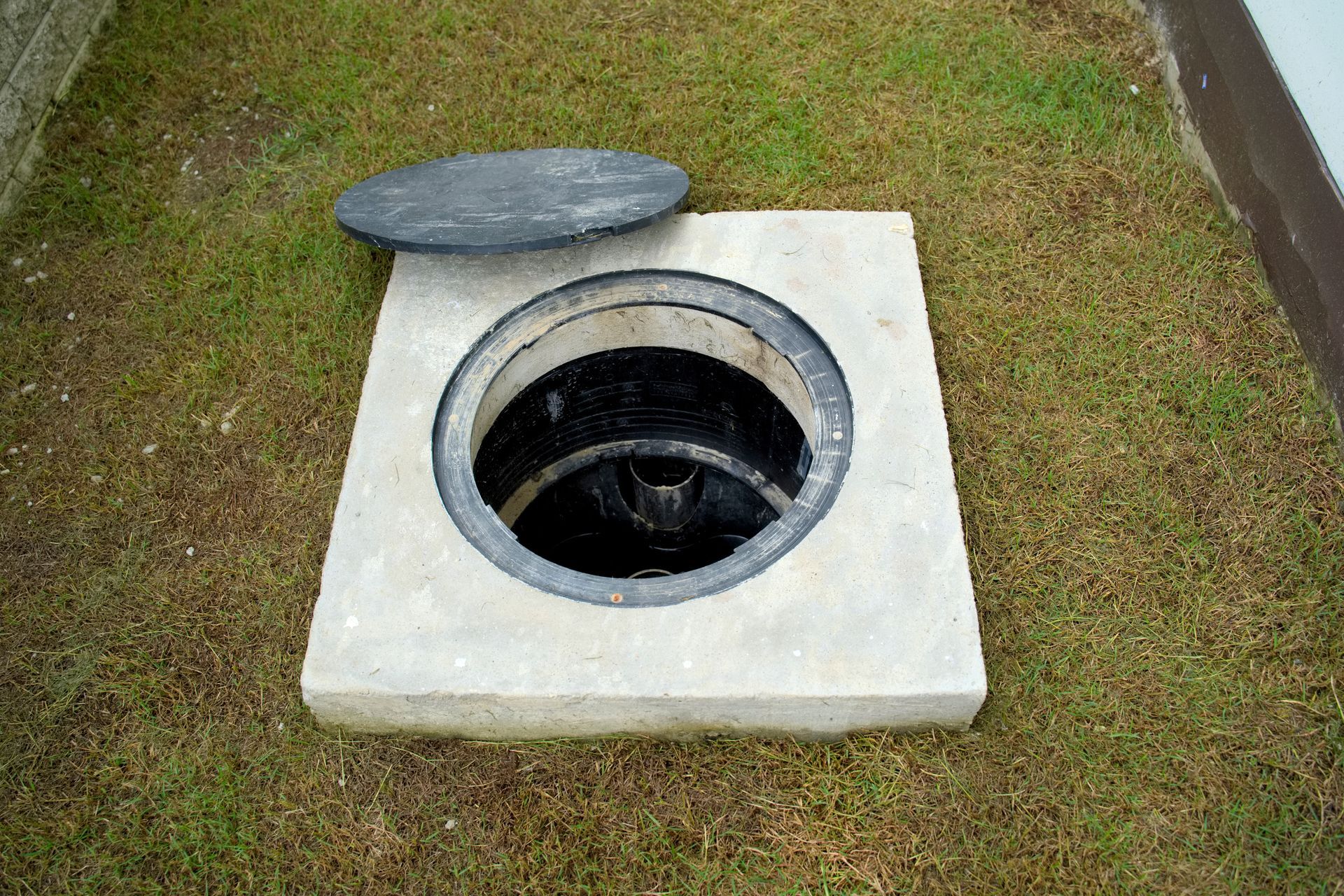 Open manhole in a concrete square, lid off, set in grass.