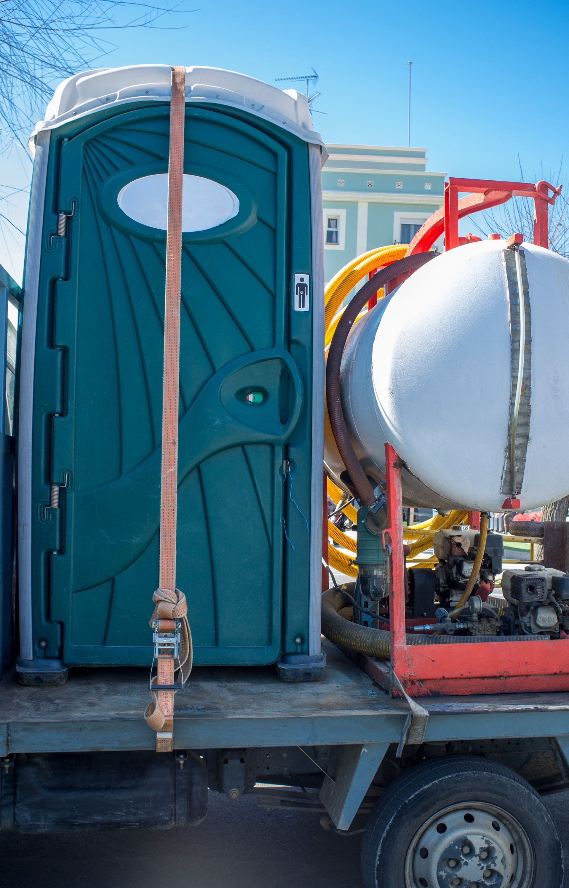 Green portable toilet secured on a truck bed with a water tank in the background. Blue sky.
