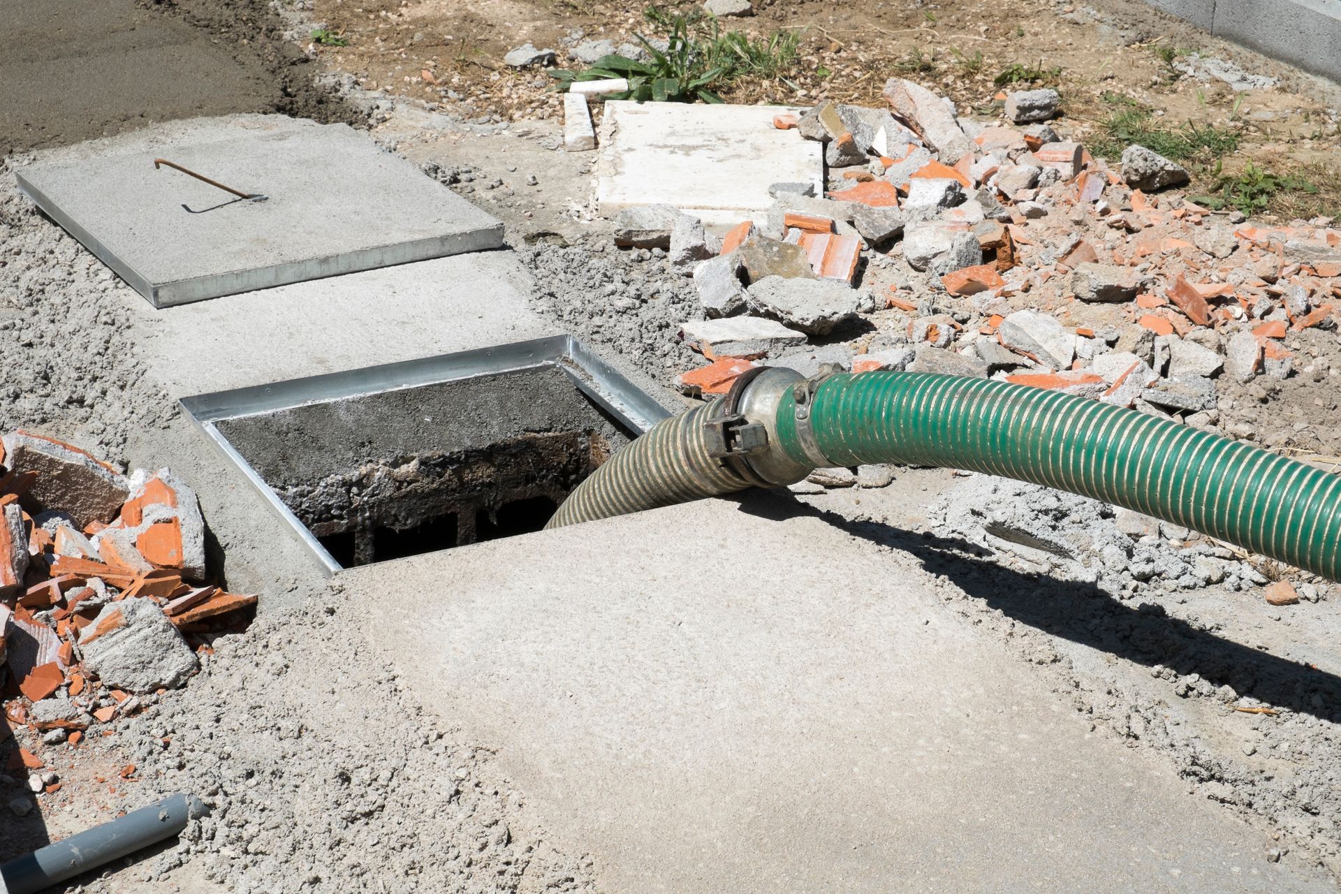 Green hose pumping liquid from a concrete septic tank lid, surrounded by construction debris.