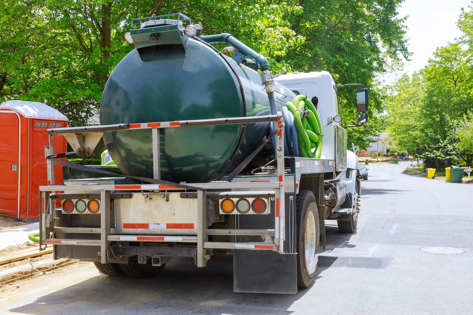 Septic tank truck with green tank parked on a street near a portable toilet and trees.