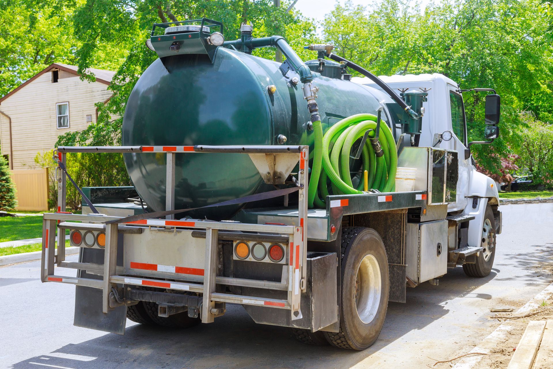Rear view of a septic tank truck, dark green tank, parked on a street near a residential neighborhood.