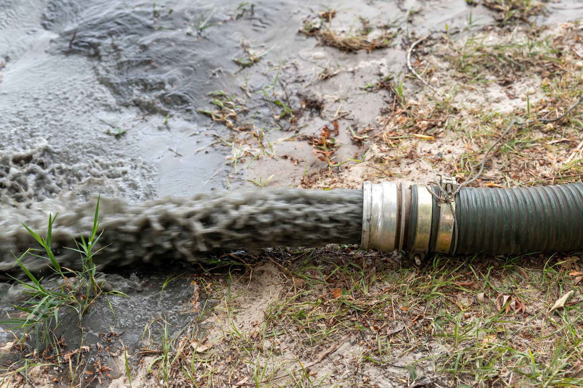 Dark liquid discharging from a hose onto muddy ground, with some grass.