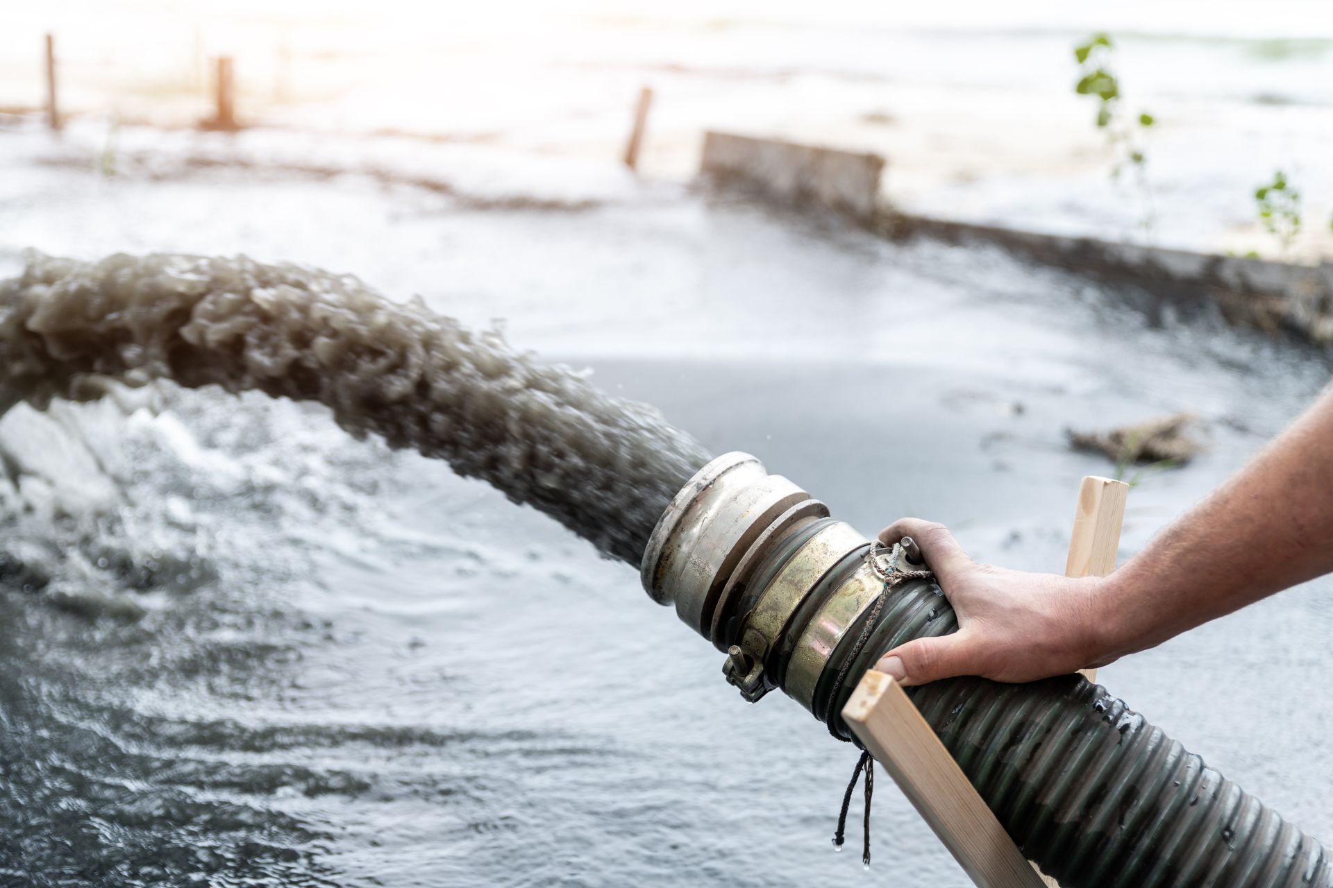 A hose expels dark, murky water into a body of water; a hand steadies the hose.