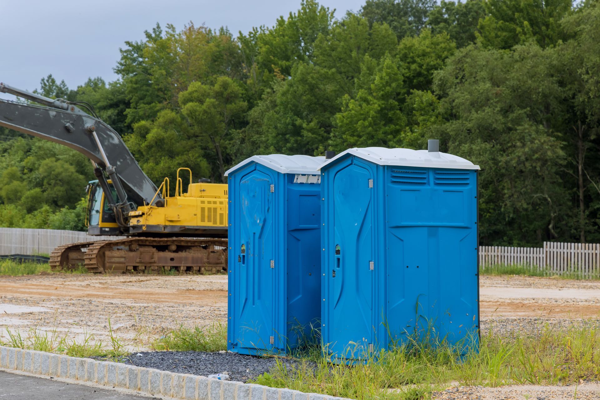 Two blue portable toilets on a gravel lot; a yellow excavator and trees are in the background.