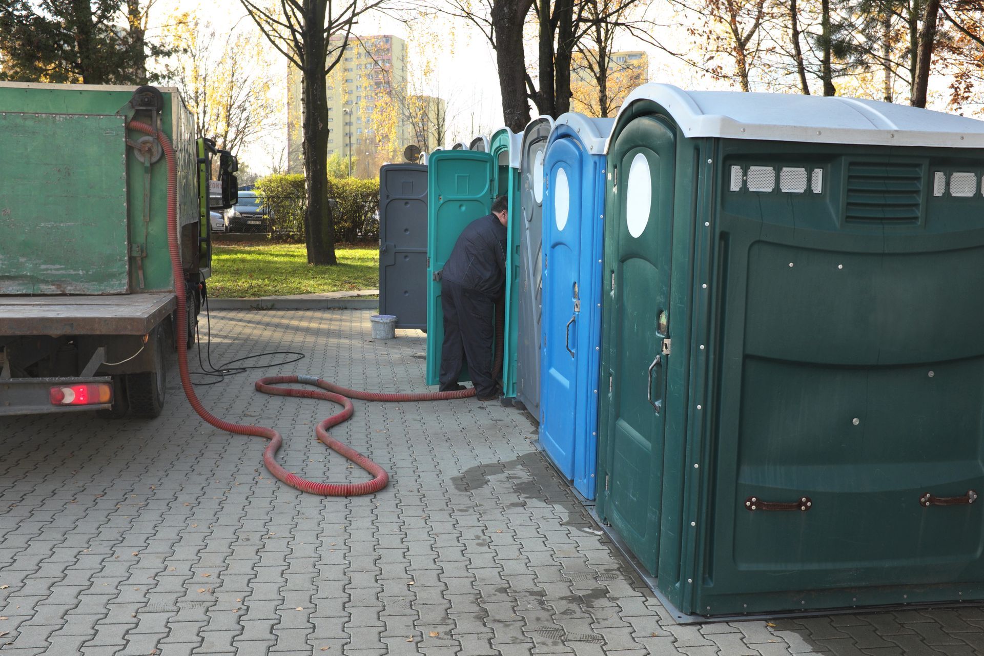 A person servicing portable toilets lined up on a paved area, hose extended from a truck.
