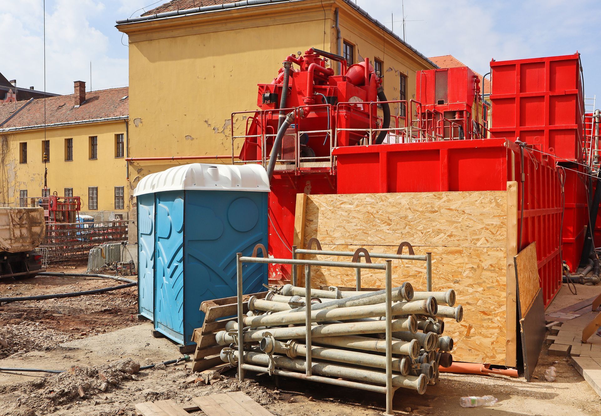 Construction site with a red machine, blue portable toilet, and building in the background.
