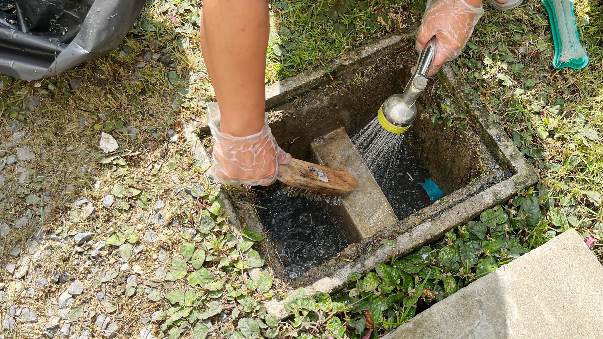 Person cleaning a concrete utility box with brush and hose, outdoors on grass.