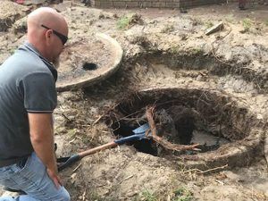Man kneels near an open septic tank. He looks inside while holding a shovel. Dirt surrounds the tank.