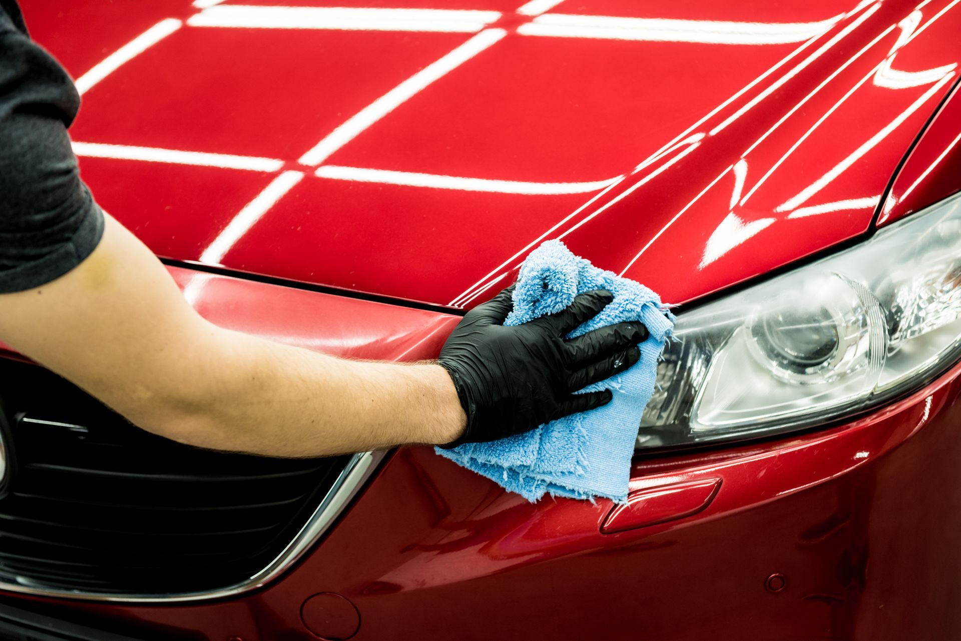 Person wearing black gloves wiping a red car's headlight with a blue microfiber cloth.