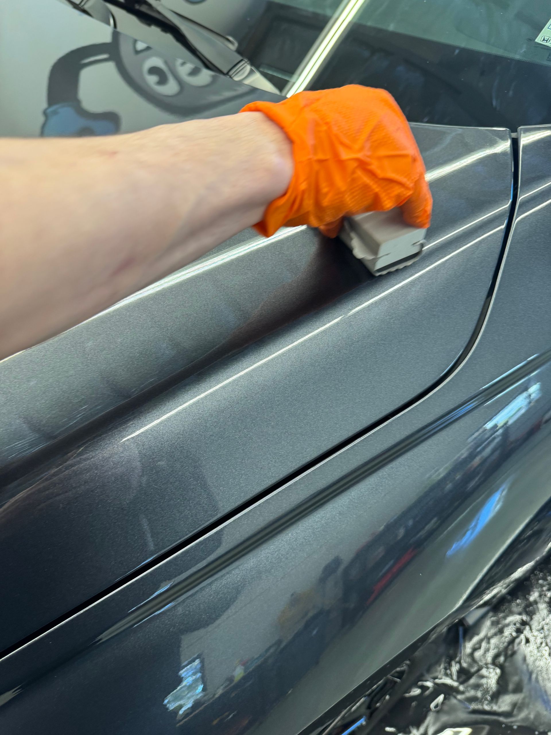Person in orange gloves applying ceramic coating to a gray car's surface.