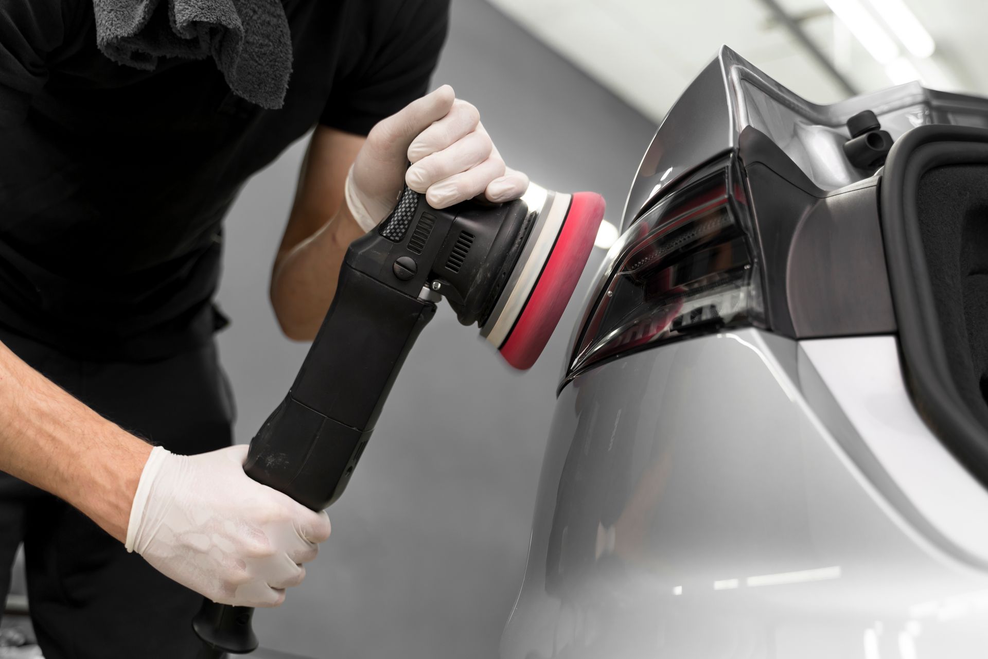 A person wearing gloves using a polisher on a silver car's taillight in a garage.