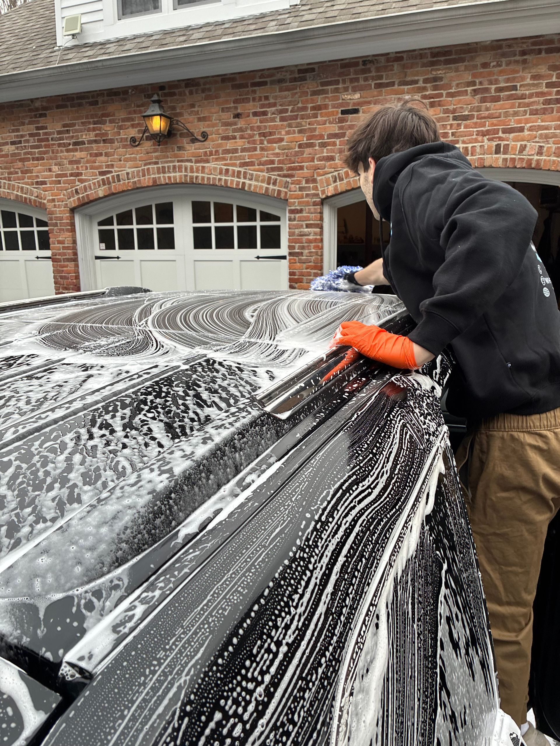 Person washing a black car with soapy water; brick garage in the background.