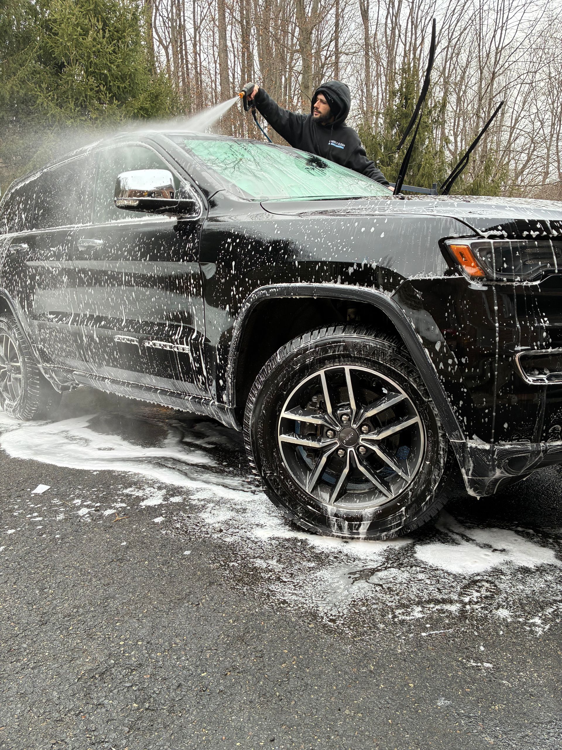 Person washing a black SUV with foamy soap outdoors on a paved surface.