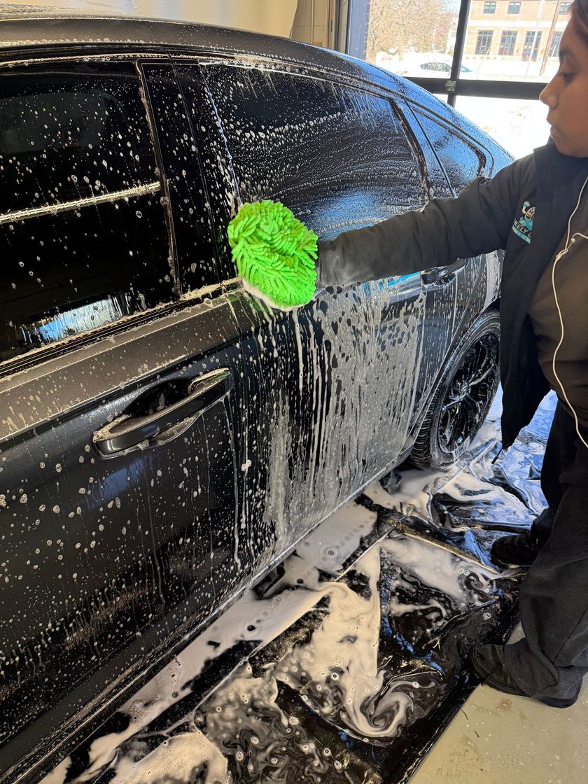 Person washing a black car with a green sponge at a car wash, covered in soapy suds.
