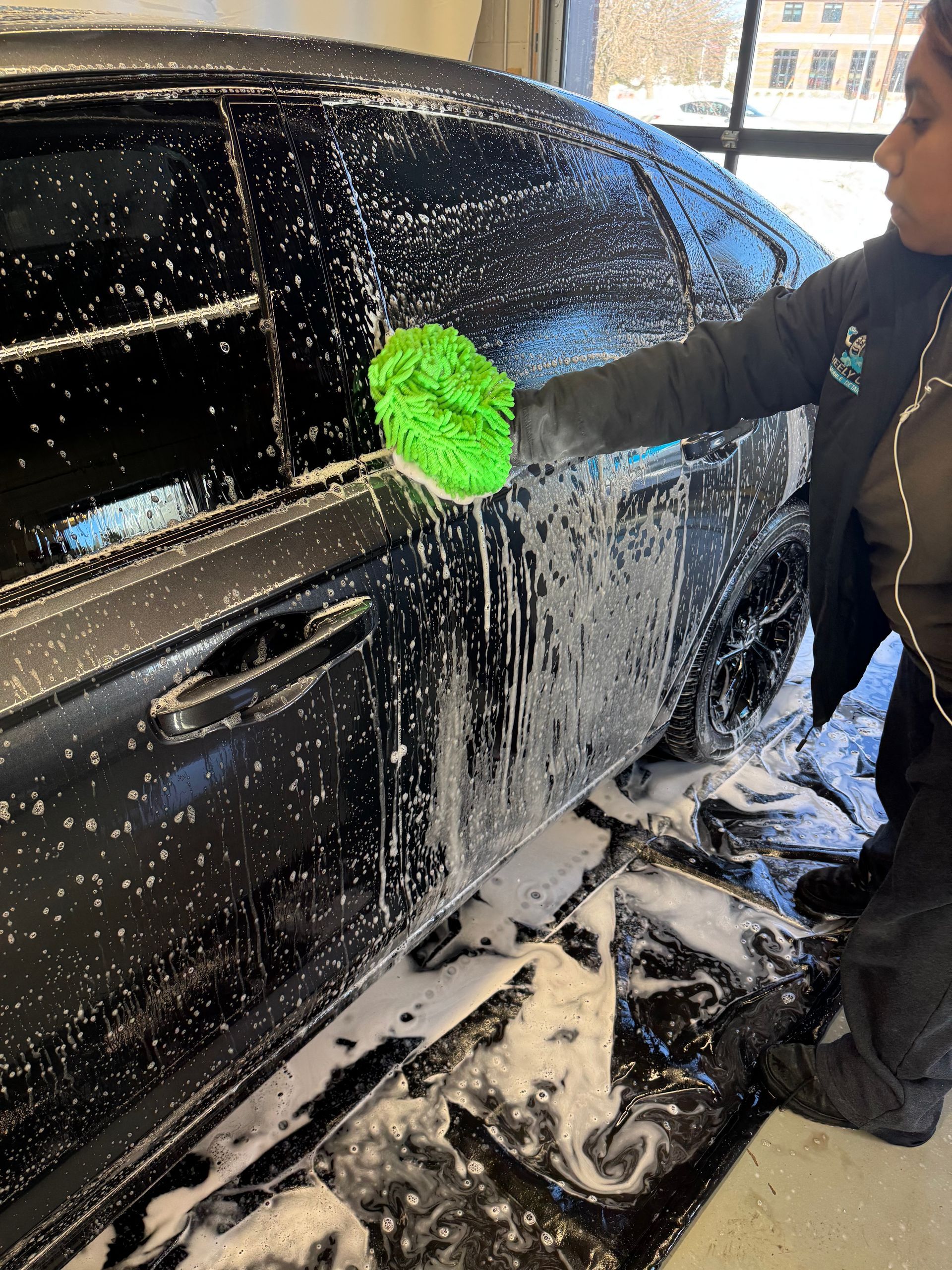 Person washing a black car with a green sponge at a car wash, covered in soapy suds.
