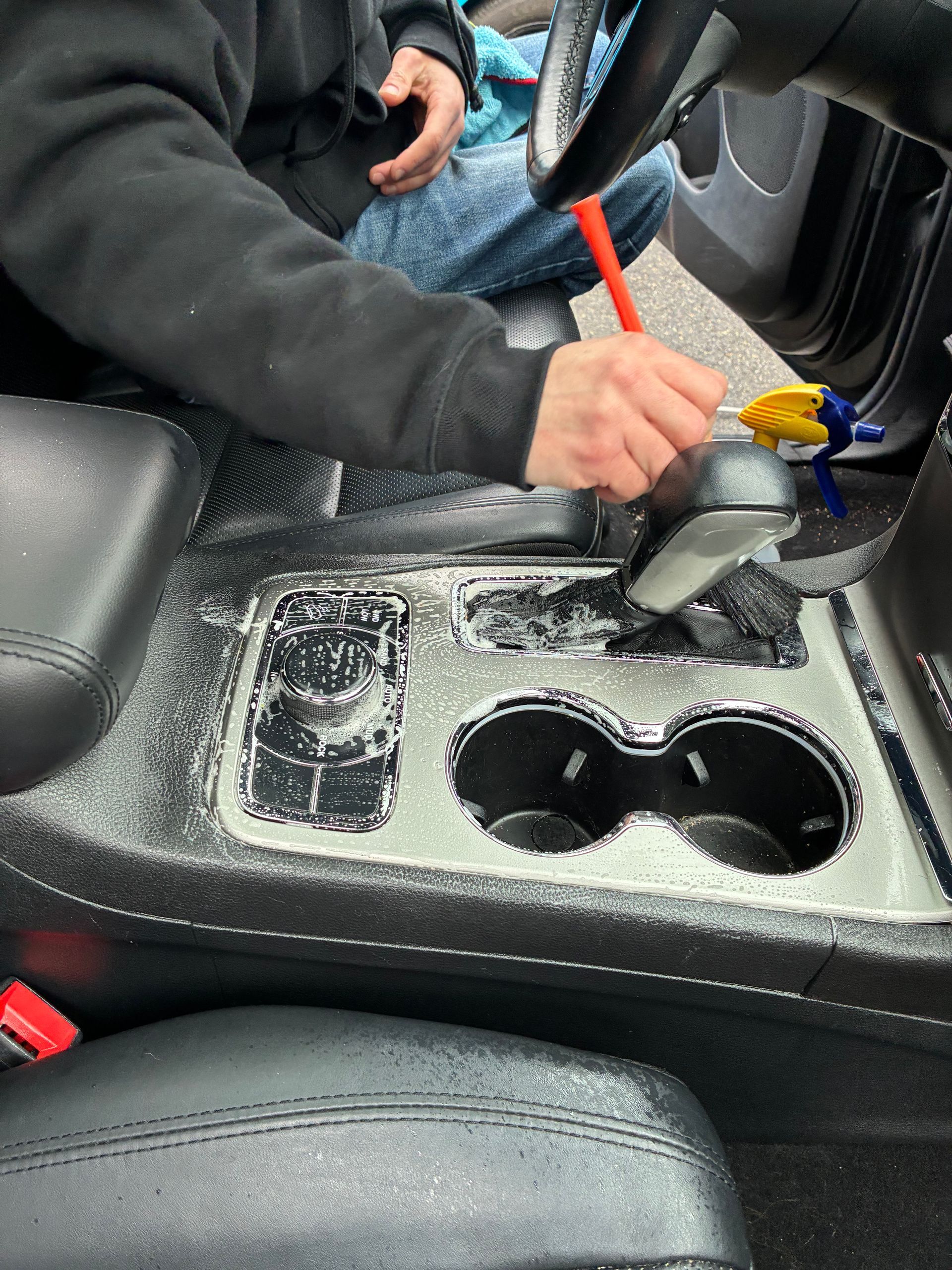 Person cleaning a car's center console with cleaning product and a brush; interior shot with black leather seats and console.