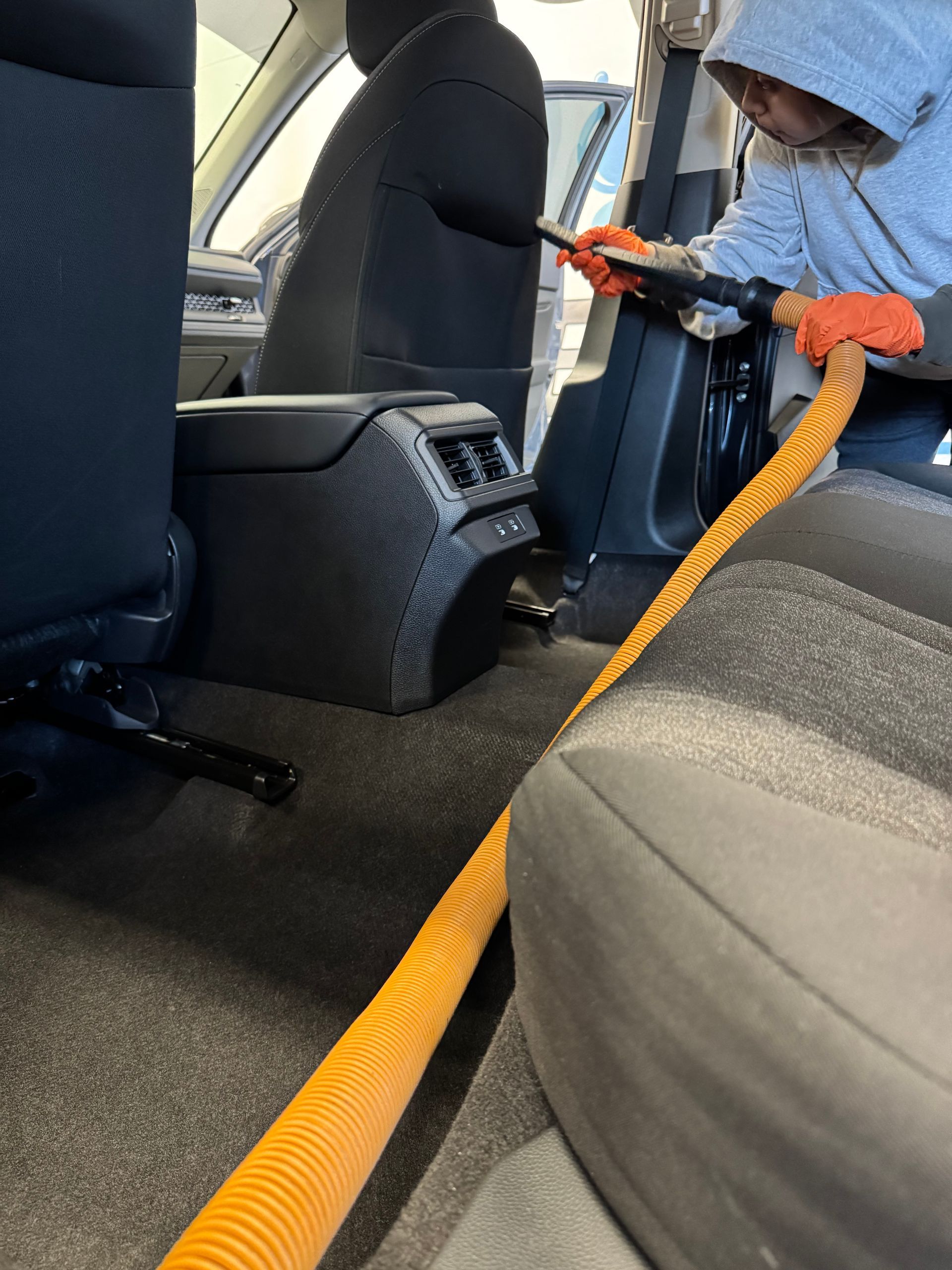 Person vacuuming the interior of a car. An orange hose extends from the vacuum to clean the seats and floor.