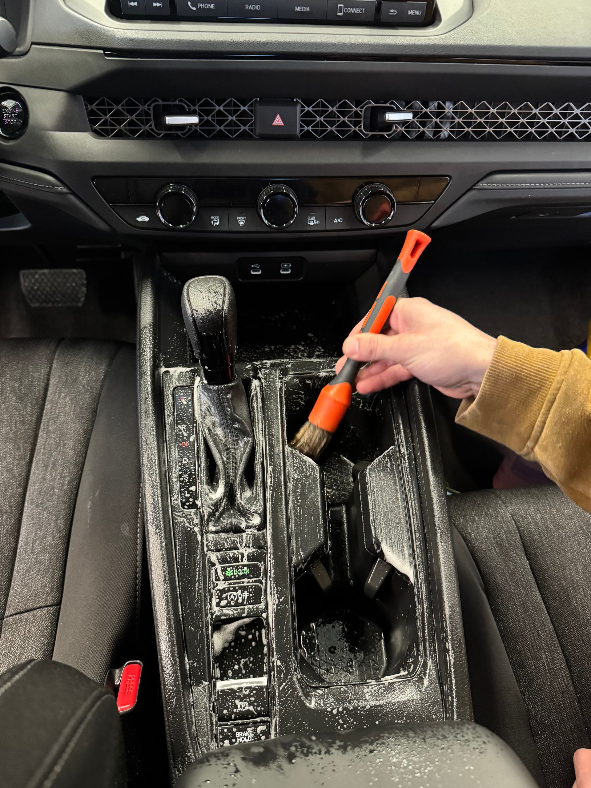A person cleaning a car's center console with a brush. Interior is black with white foam cleaner.