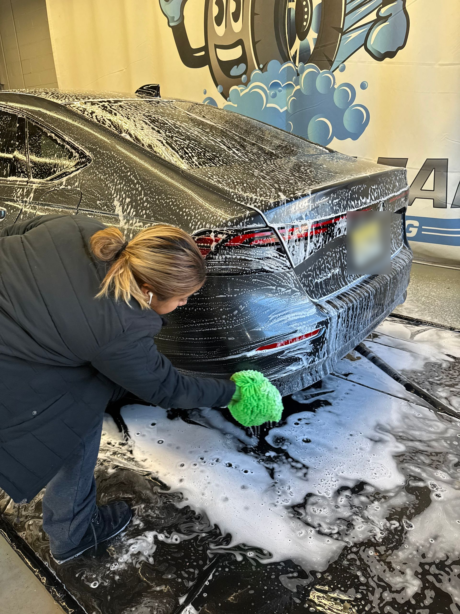 Person washing a black car at a car wash. They are covered in soap suds, using a green wash mitt.