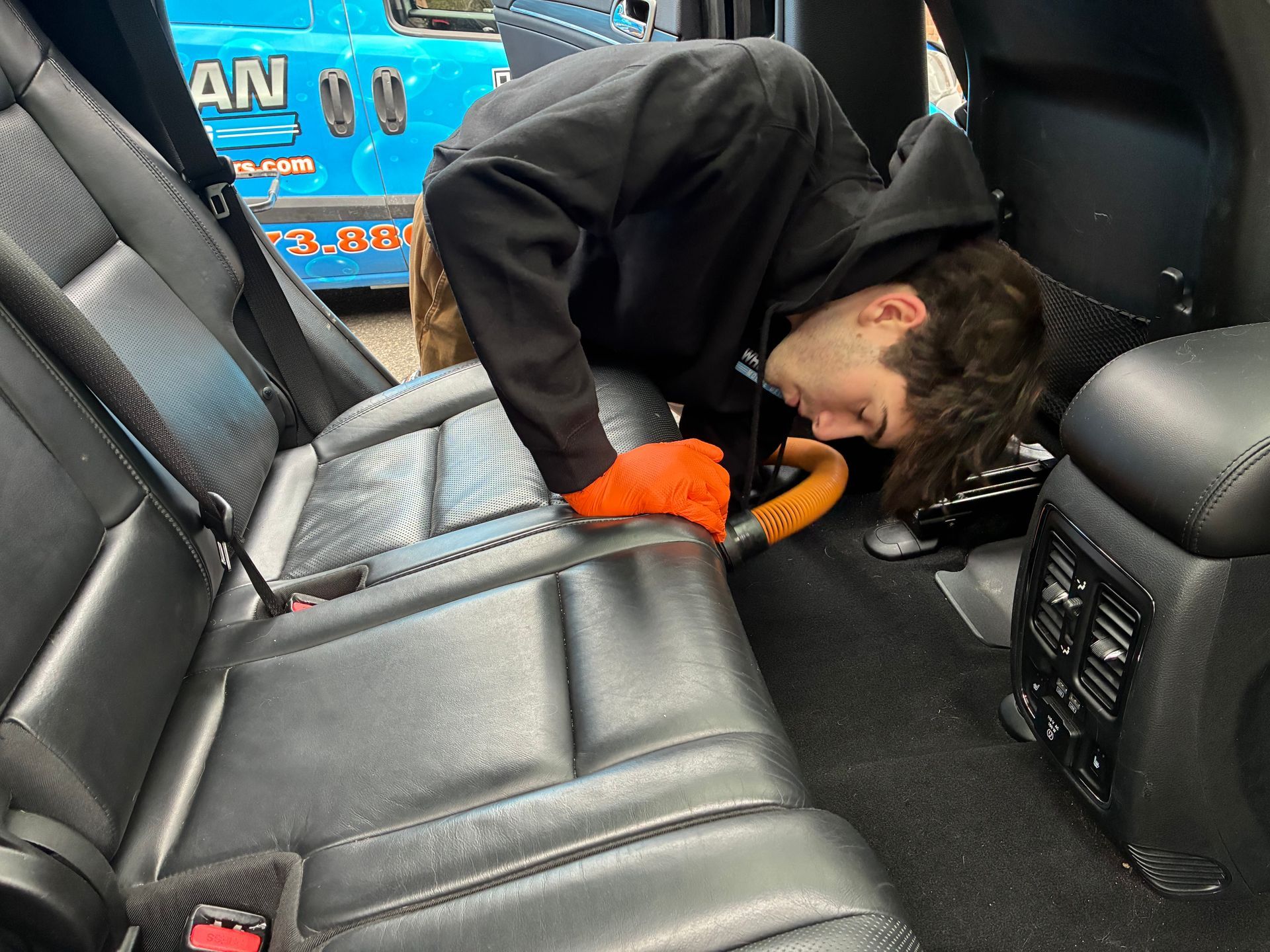 A person vacuums the back seat of a black car, wearing orange gloves.