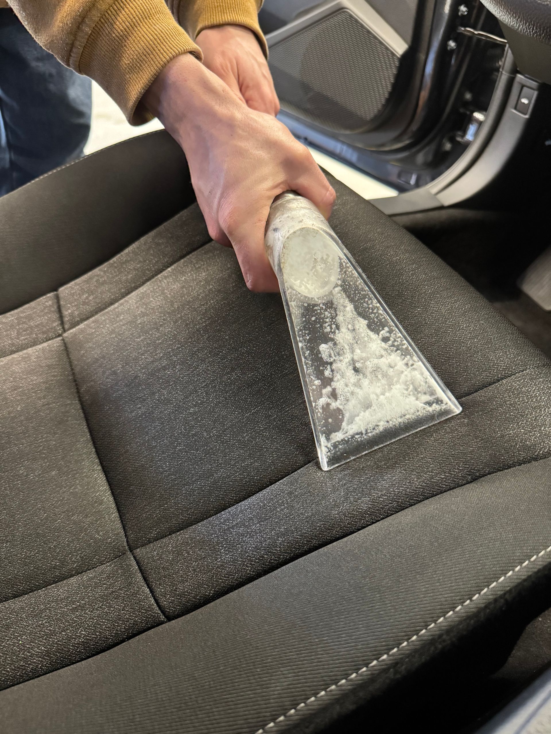 Person vacuuming a car seat with a handheld cleaning tool, showing cleaning solution.