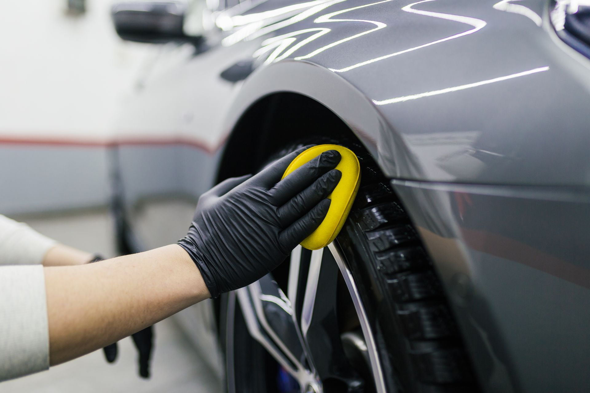 Person wearing black gloves cleaning a gray car tire with a yellow sponge.