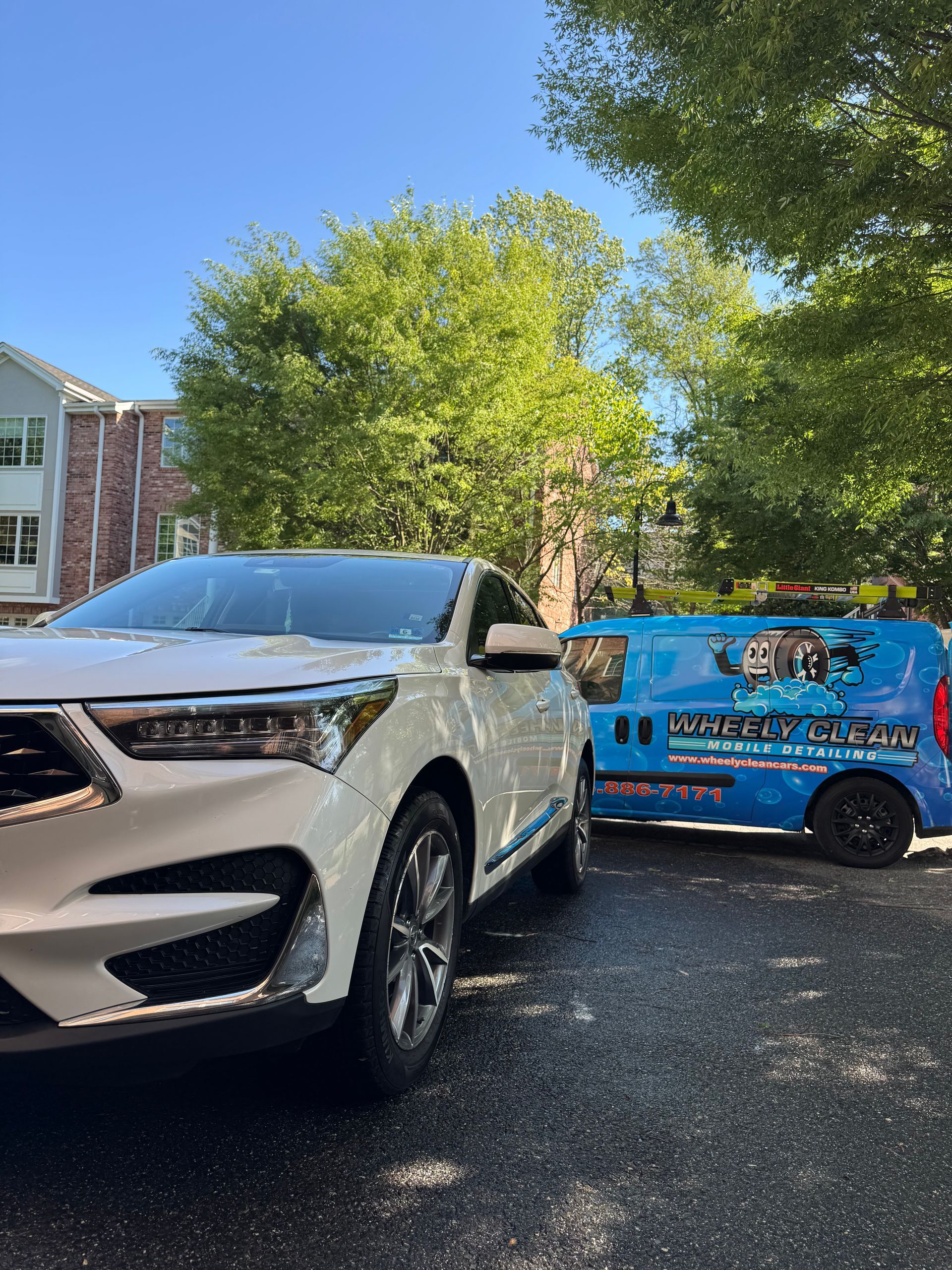 White SUV next to a blue service van parked on a street in front of a building and trees.
