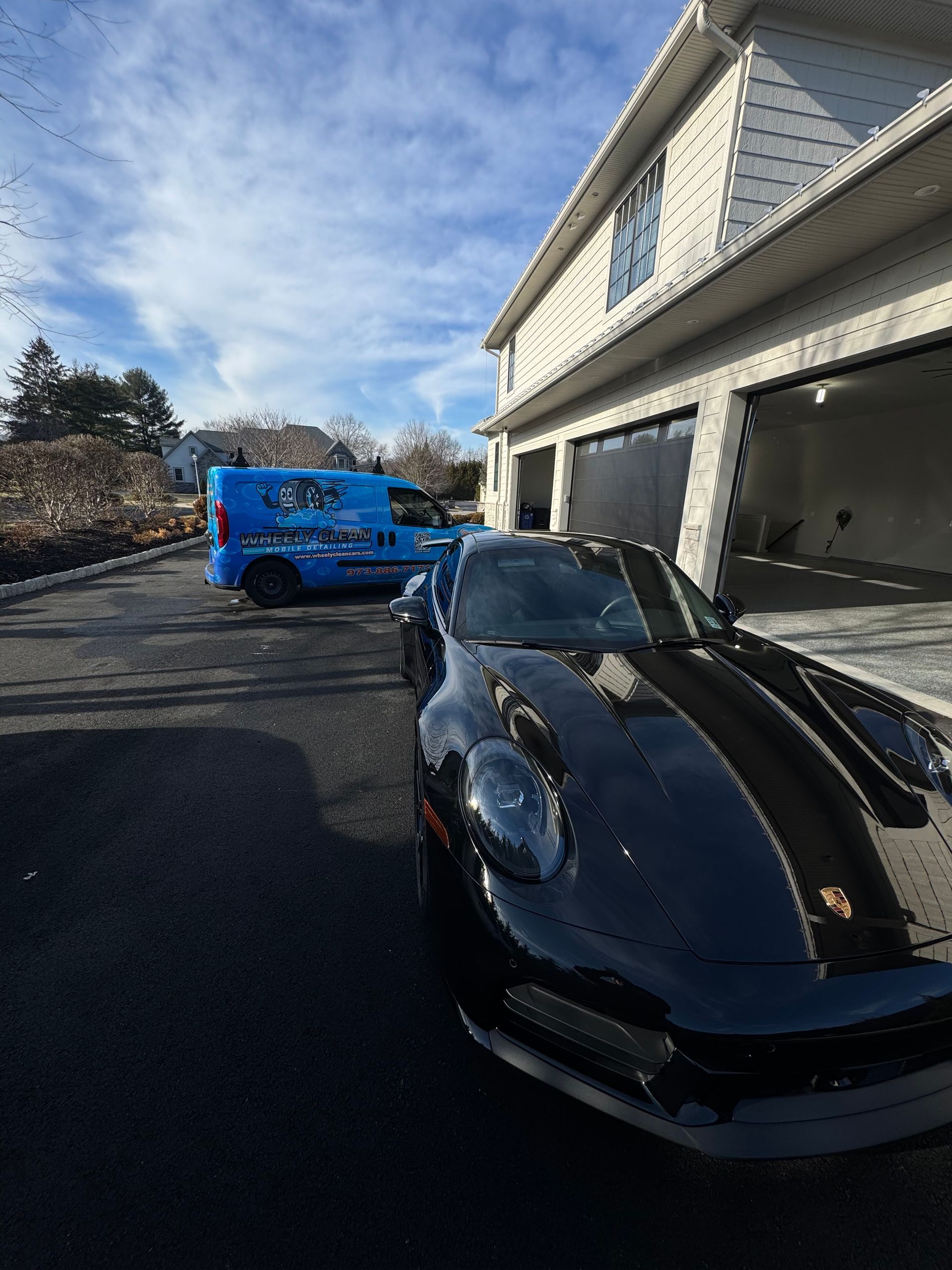 Black Porsche parked in front of a house with a garage; a blue van is in the background.