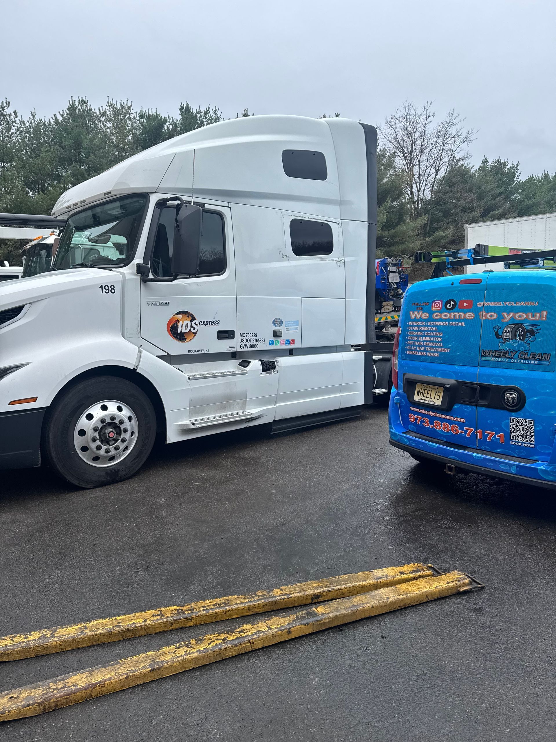 White semi-truck parked next to a blue vehicle; both are on a wet parking lot. Yellow barrier in the foreground.