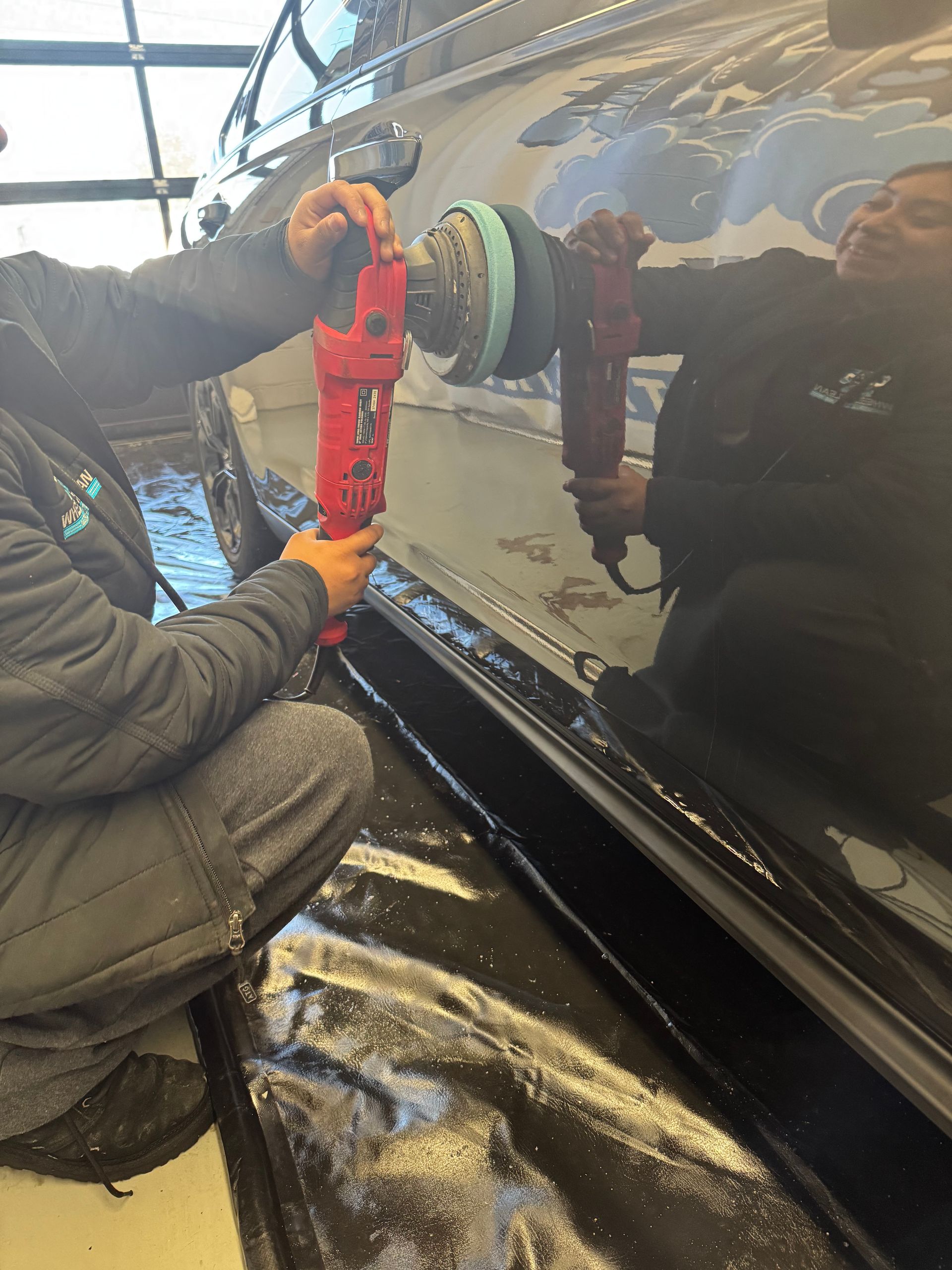 Person using a red buffer tool on a black car, in a detailing shop.
