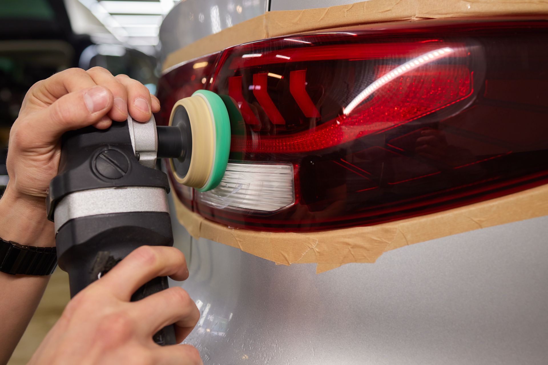 Person using a polishing tool on a car tail light, taped off to protect the car's body.