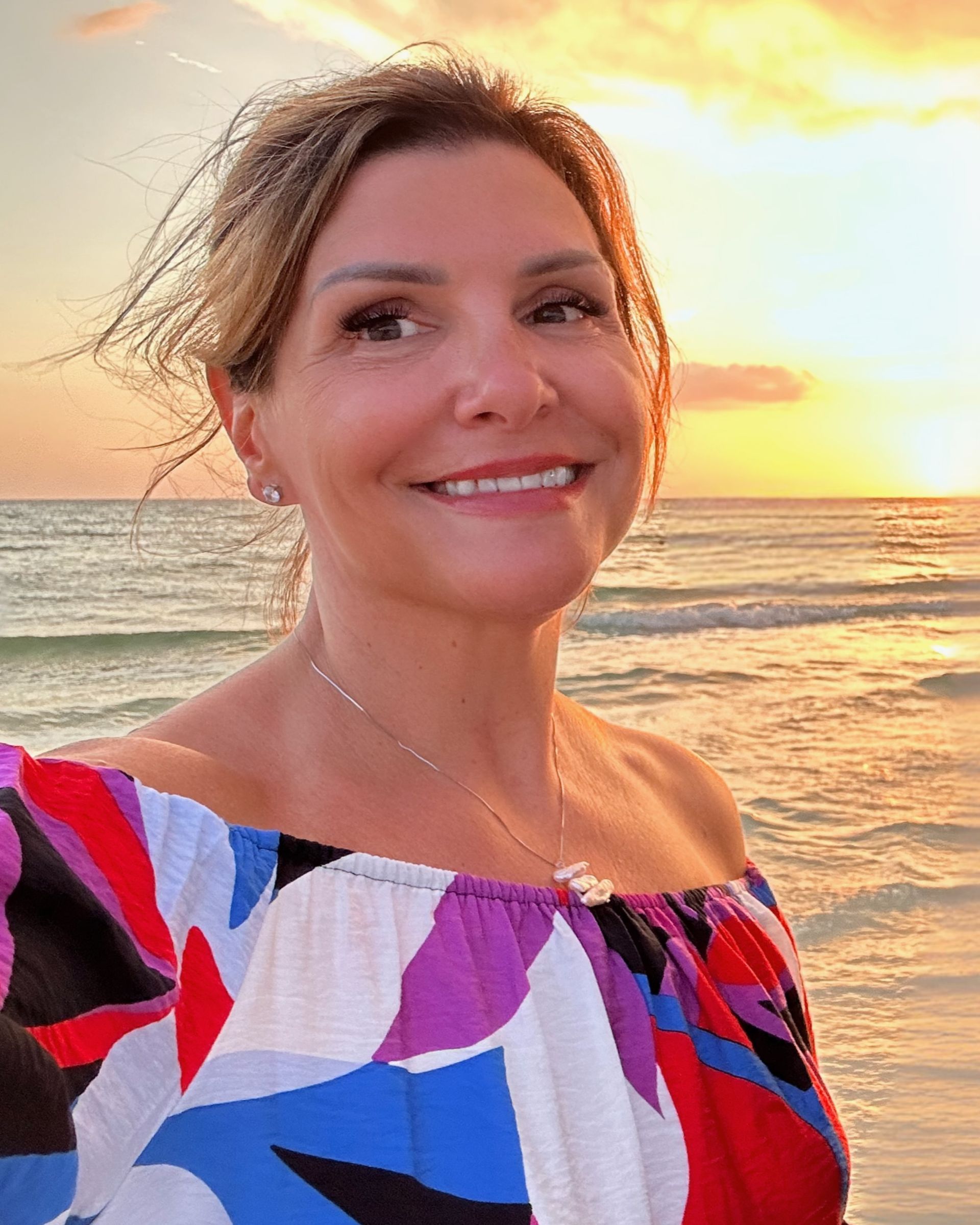 A smiling woman stands on a beach at sunset wearing a colorful off-the-shoulder top.