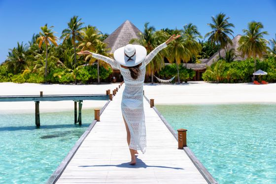 A person in a white lace dress and wide-brimmed hat walks with arms outstretched on a pier toward a tropical beach.