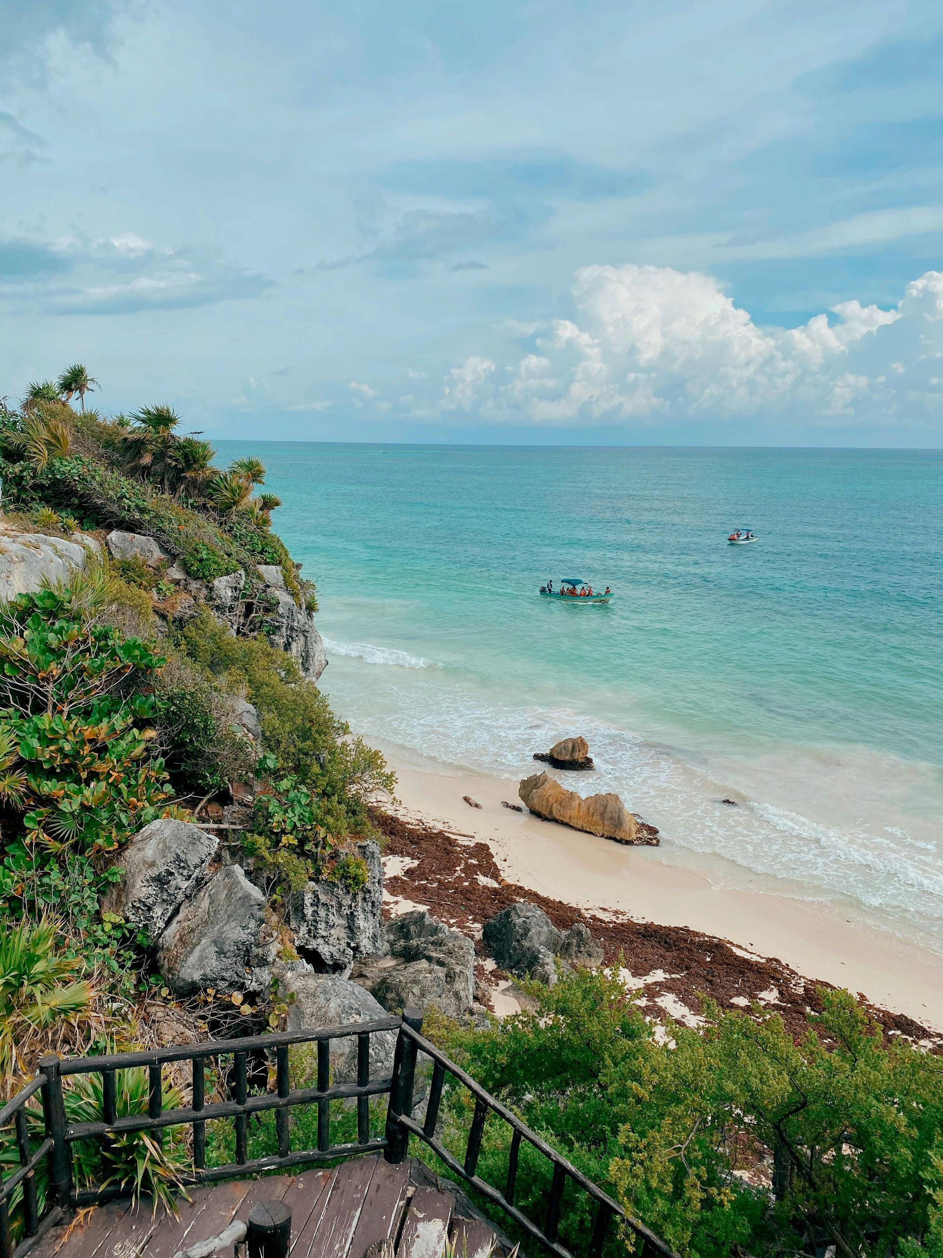 Clifftop view of turquoise ocean and sandy beach with rocks and boats under a cloudy sky.