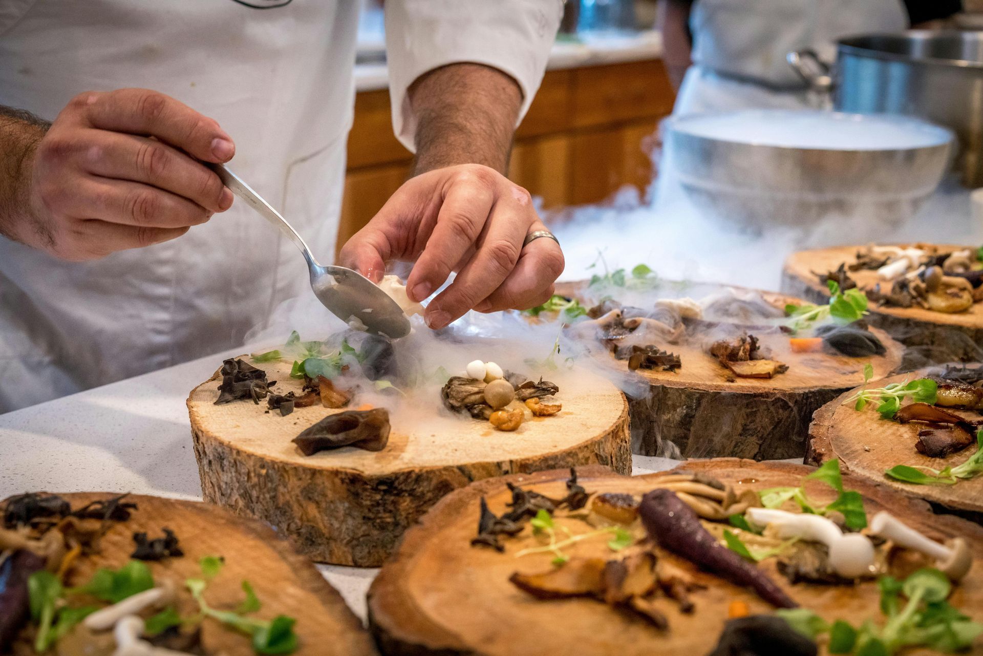 Chef spoons something onto a wood-plank dish, surrounded by other mushroom dishes with dry ice.