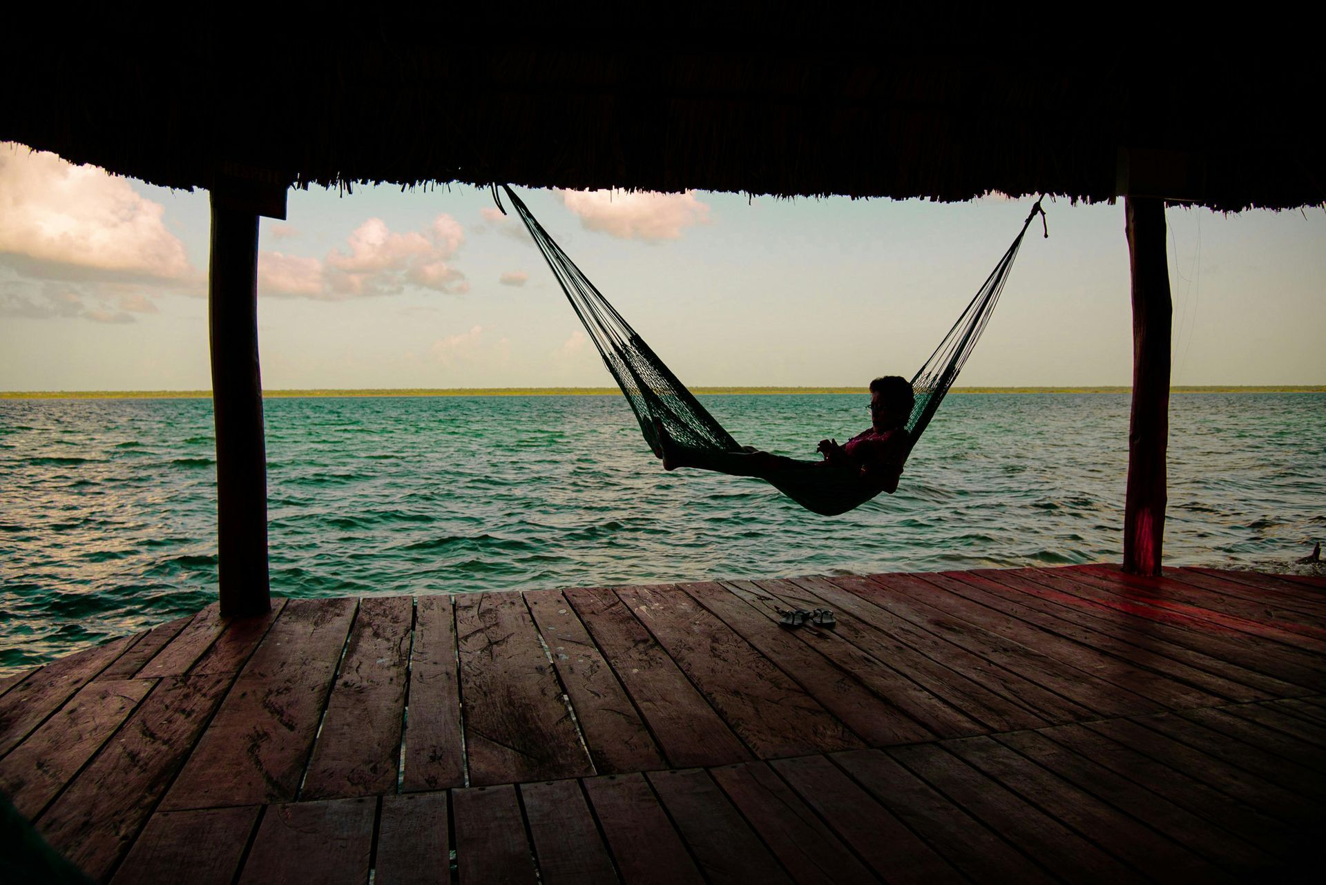 Person relaxes in a hammock suspended over ocean on a wooden deck.